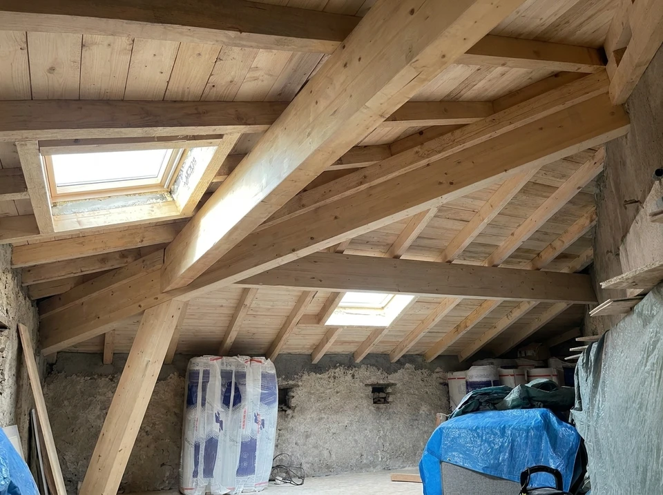 Photographie d'une charpente en ossature bois robuste vue en gros plan ou sous angle dynamique, montrant des poutres massives assemblées avec des techniques traditionnelles (mortaises, tenons, chevilles bois), dans une ambiance de chantier ou de maison en rénovation. La lumière naturelle doit mettre en évidence la qualité et la structure fiable du bois. Pas de personne, pas de texte, pas de logo.